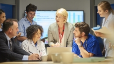 Several people standing and sitting around a table with a screen in the background that has a graph on it Several people standing and sitting around a table with a screen in the background that has a graph on it