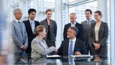 Several business people standing around a table watching two people sitting down shaking hands Several business people standing around a table watching two people sitting down shaking hands