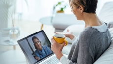 Person sitting on a couch while holding a cup and talking to a healthcare provider on their computer Person sitting on a couch while holding a cup and talking to a healthcare provider on their computer