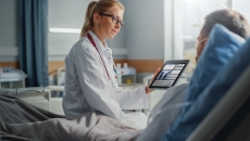 Healthcare provider sitting on a patient's hospital bed while holding a tablet showing diagnostic images of lungs Healthcare provider sitting on a patient's hospital bed while holding a tablet showing diagnostic images of lungs