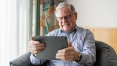 Person sitting in a chair holding a tablet while smiling and wearing glasses Person sitting in a chair holding a tablet while smiling and wearing glasses