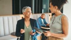Two people standing in a hallway in a building with one person sitting down behind them Two people standing in a hallway in a building with one person sitting down behind them