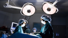 Healthcare providers in an operating room looking down at a patient on a table with large lights above them Healthcare providers in an operating room looking down at a patient on a table with large lights above them