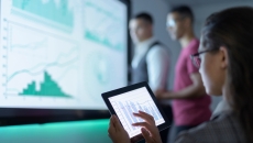 Two people standing in front of a screen looking at a graph and one person sitting down looking at a tablet Two people standing in front of a screen looking at a graph and one person sitting down looking at a tablet