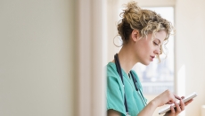 Healthcare provider standing in a hallway wearing green scrubs and a stethoscope around their neck while looking at a tablet Healthcare provider standing in a hallway wearing green scrubs and a stethoscope around their neck while looking at a tablet