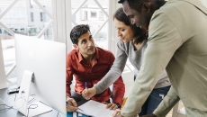 Person in a red shirt sitting at a desk in front of a computer with two people standing beside him looking at the computer Person in a red shirt sitting at a desk in front of a computer with two people standing beside him looking at the computer