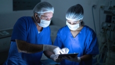 Two people wearing scrubs and masks in a procedure room look at a tablet. Two people wearing scrubs and masks in a procedure room look at a tablet.