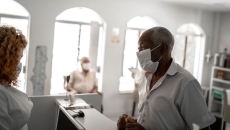 An older patient wearing a mask checking in a with a receptionist An older patient wearing a mask checking in a with a receptionist