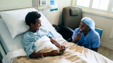 Healthcare provider sitting next to a hospital bed with a teenager in it looking at a phone