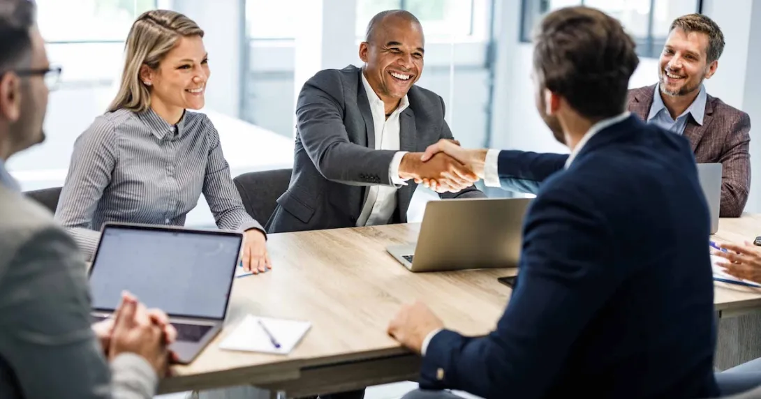 Two people sitting across from each other at a table and shaking hands while other watch Two people sitting across from each other at a table and shaking hands while other watch