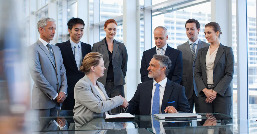 Several business people standing around a table watching two people sitting down shaking hands