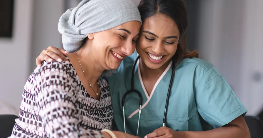 A healthcare provider with scrubs on sitting next to an oncology patient wearing a head scarf A healthcare provider with scrubs on sitting next to an oncology patient wearing a head scarf