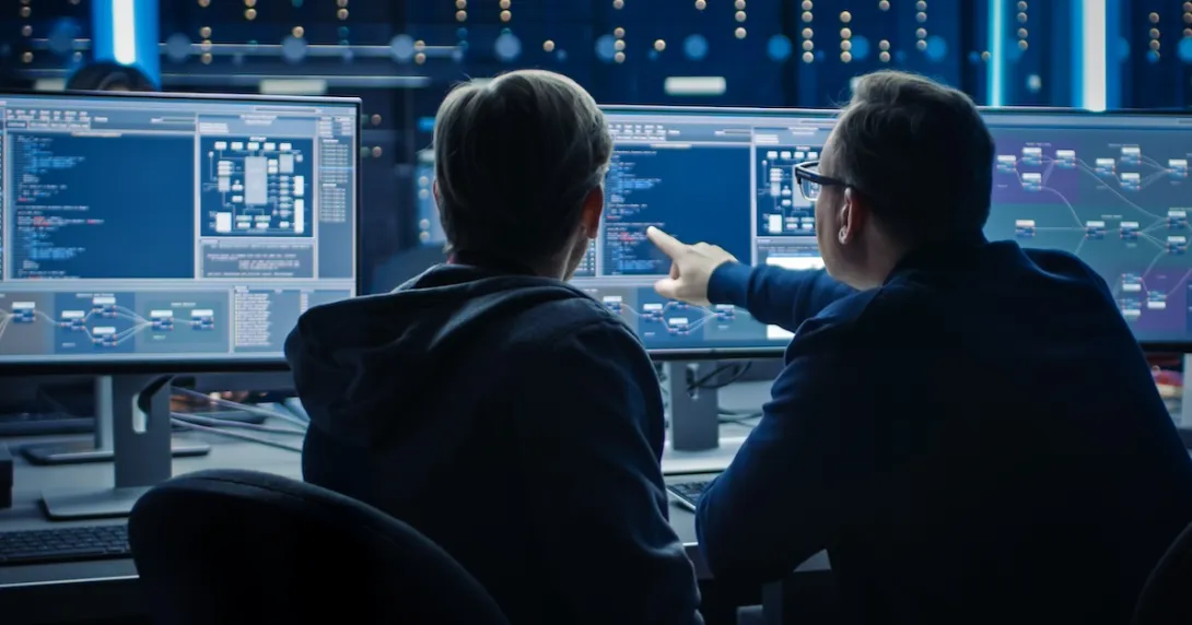 Two individuals sitting in front of two computer monitors with their backs turned toward the camera Two individuals sitting in front of two computer monitors with their backs turned toward the camera