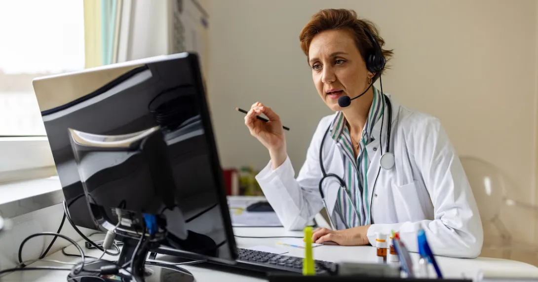 Healthcare provider sitting at a desk wearing a lab coat and headphones while talking to someone on a computer Healthcare provider sitting at a desk wearing a lab coat and headphones while talking to someone on a computer