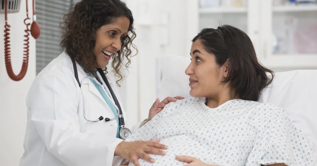 Healthcare worker standing beside a pregnant person lying in bed while both are touching the pregnant person's stomach Healthcare worker standing beside a pregnant person lying in bed while both are touching the pregnant person's stomach