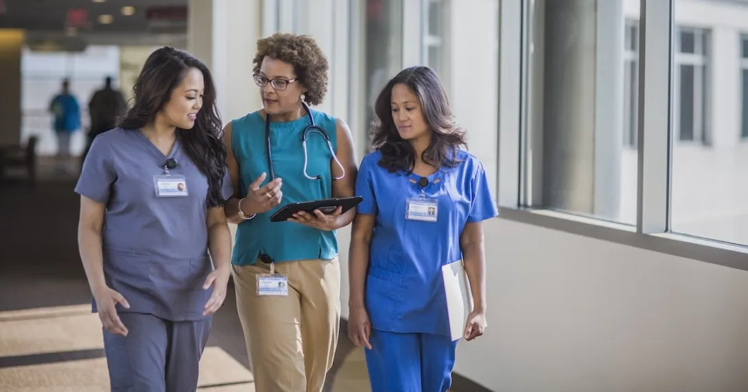 Three healthcare providers walking through a hall while looking at a tablet Three healthcare providers walking through a hall while looking at a tablet