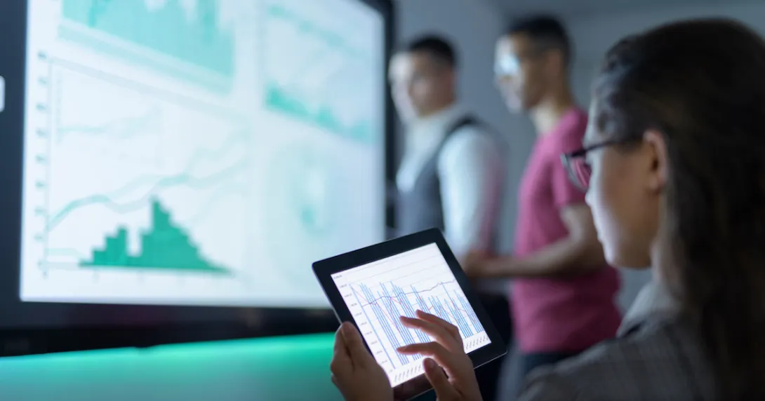Person sitting while looking at a tablet with two other people standing up looking at a graph on a screen Person sitting while looking at a tablet with two other people standing up looking at a graph on a screen