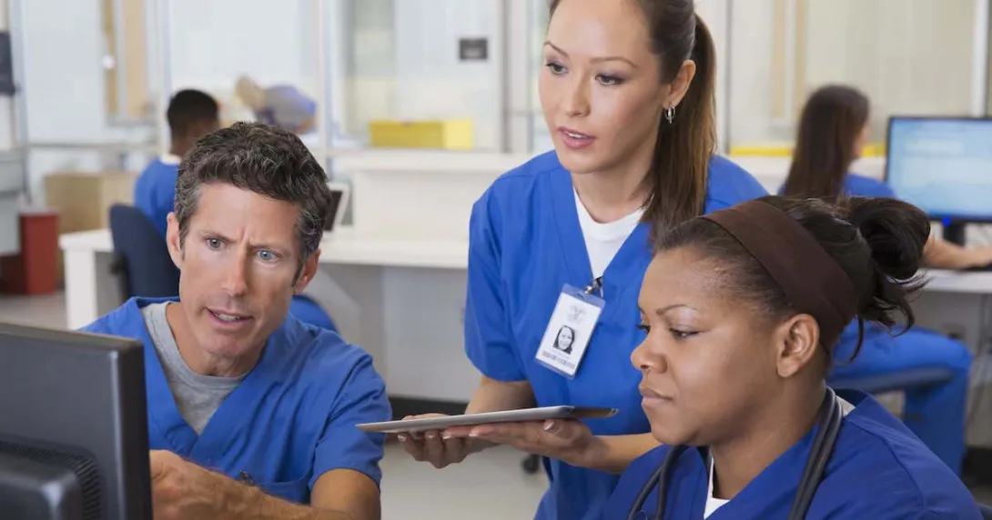 Three healthcare providers wearing scrubs sitting around a computer looking at the screen Three healthcare providers wearing scrubs sitting around a computer looking at the screen