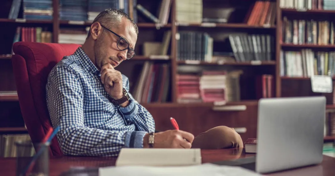 Person sitting at a desk writing something down on a piece of paper with a bookshelf behind them Person sitting at a desk writing something down on a piece of paper with a bookshelf behind them