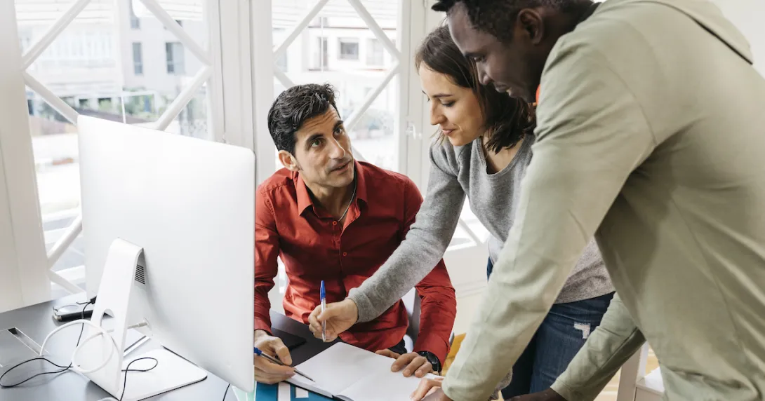 Person in a red shirt sitting at a desk in front of a computer with two people standing beside him looking at the computer Person in a red shirt sitting at a desk in front of a computer with two people standing beside him looking at the computer