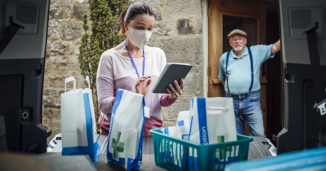 A woman using a tablet delivering medications while a man stands in his doorway in the background A woman using a tablet delivering medications while a man stands in his doorway in the background