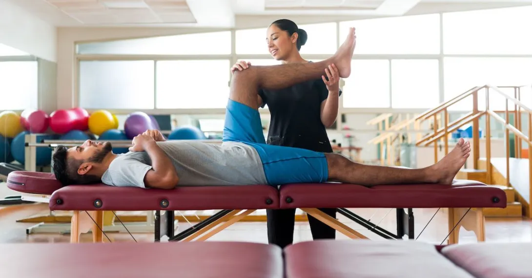 A male patient lying on his back on a massage table and a female physical therapist holding his leg up. A male patient lying on his back on a massage table and a female physical therapist holding his leg up.