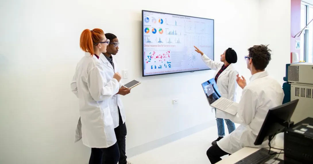 Professional team of medical scientists in a laboratory gathered around a wall monitor discussing charts and data Professional team of medical scientists in a laboratory gathered around a wall monitor discussing charts and data
