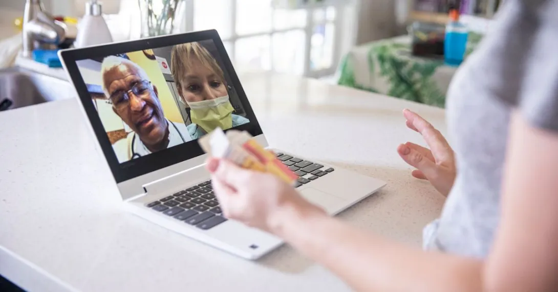 A close up of a woman holding a pill bottle while talking to two providers on her laptop. A close up of a woman holding a pill bottle while talking to two providers on her laptop.