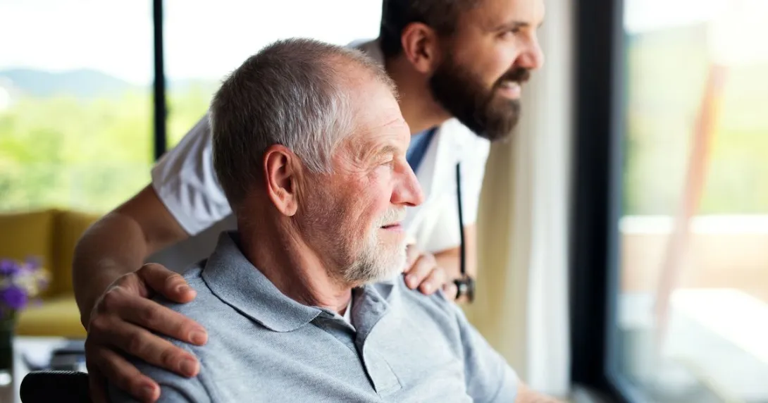 A person in a wheelchair and a healthcare provider standing beside them looking out a window A person in a wheelchair and a healthcare provider standing beside them looking out a window