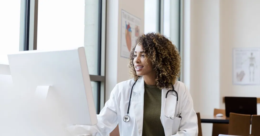 Healthcare provider with a lab coat on sitting at a desk and looking at a computer Healthcare provider with a lab coat on sitting at a desk and looking at a computer