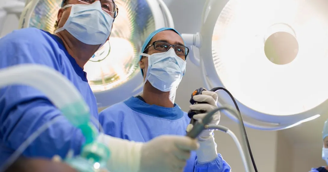 Two surgeons looking down at a camera with scrubs on while holding surgical tools Two surgeons looking down at a camera with scrubs on while holding surgical tools