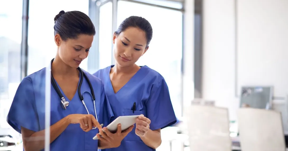 Two healthcare providers in scrubs standing side-by-side looking at a tablet Two healthcare providers in scrubs standing side-by-side looking at a tablet
