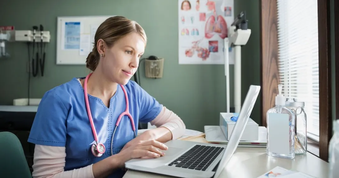 Healthcare provider in scrubs sitting at a desk and looking at a computer Healthcare provider in scrubs sitting at a desk and looking at a computer