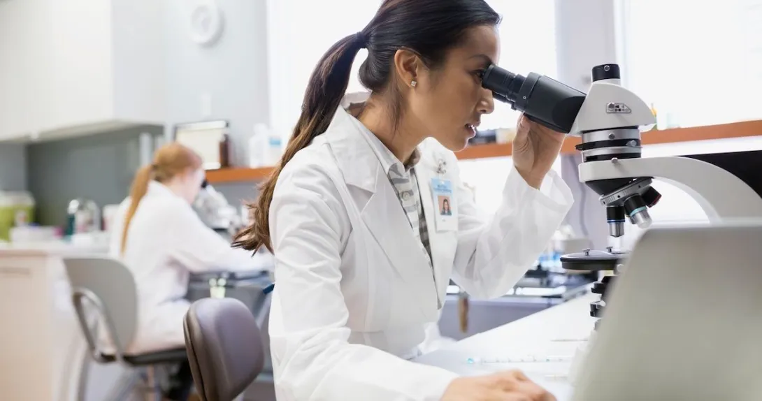 Person sitting down looking into a microscope while wearing a lab coat Person sitting down looking into a microscope while wearing a lab coat