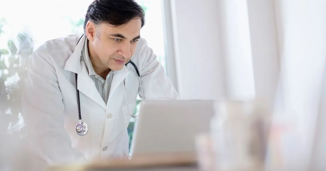 Healthcare provider wearing a lab coat with a stethoscope around their neck while looking at a computer Healthcare provider wearing a lab coat with a stethoscope around their neck while looking at a computer