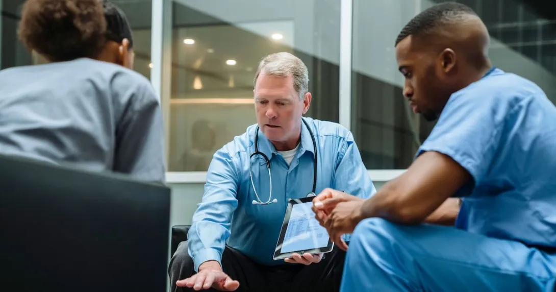 Three healthcare providers sitting around a table looking at a tablet Three healthcare providers sitting around a table looking at a tablet