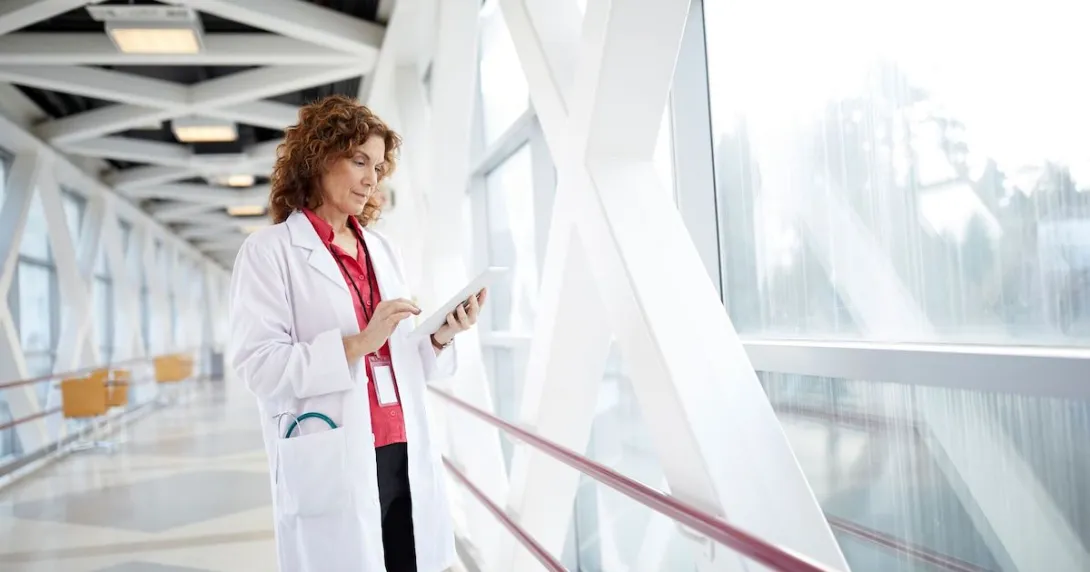 Healthcare provider standing in a hallway looking at a tablet next to a giant window Healthcare provider standing in a hallway looking at a tablet next to a giant window