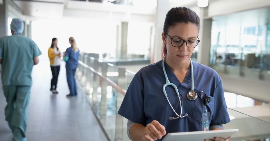 Healthcare provider looking at a tablet in the hallway of a large building Healthcare provider looking at a tablet in the hallway of a large building
