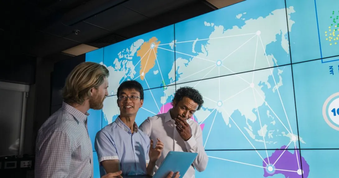 Three people standing in front of a large board showing the world map Three people standing in front of a large board showing the world map