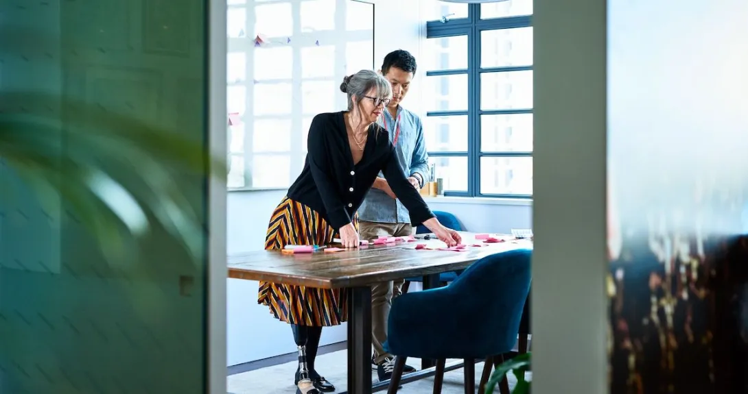 Two people standing in a room looking at something on a desk while standing above it Two people standing in a room looking at something on a desk while standing above it