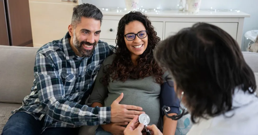 Two people sitting on a couch with one rubbing the other's stomach while both speak to a healthcare provider Two people sitting on a couch with one rubbing the other's stomach while both speak to a healthcare provider