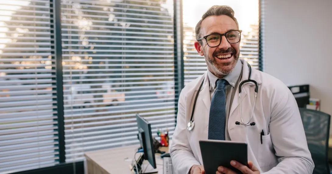Healthcare provider sitting on a desk holding a tablet while smiling Healthcare provider sitting on a desk holding a tablet while smiling