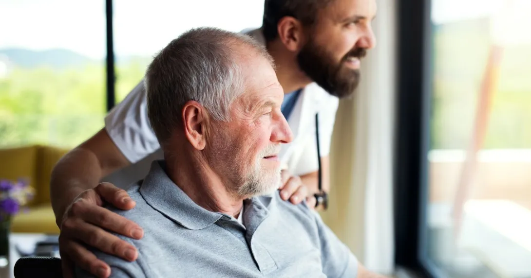 One person sitting in a chair while another person stands beside them as both look out of a window One person sitting in a chair while another person stands beside them as both look out of a window