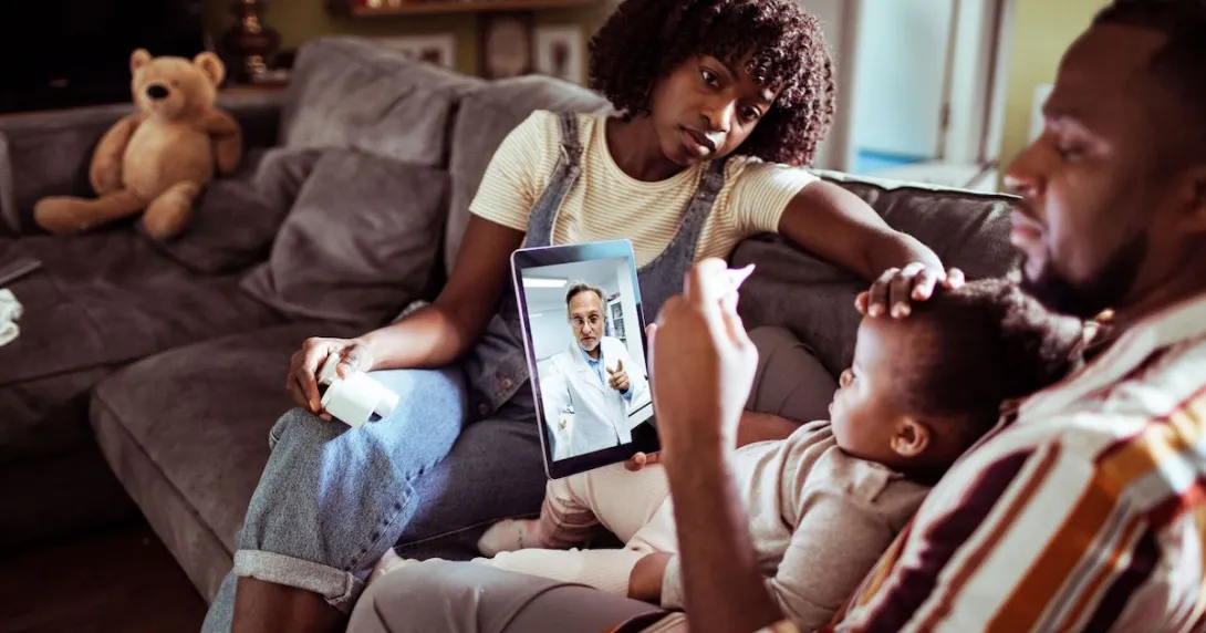 Two people sitting on a couch with a child sitting on one's lap while talking to a healthcare provider on a computer Two people sitting on a couch with a child sitting on one's lap while talking to a healthcare provider on a computer