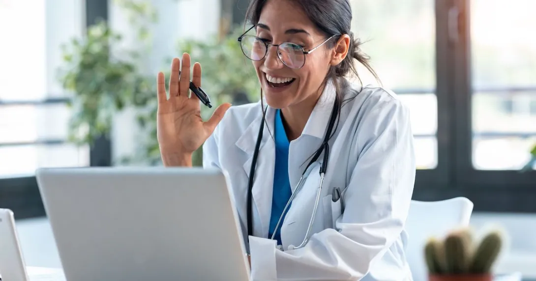 Healthcare provider sitting at a desk and looking at a computer while waving to the screen Healthcare provider sitting at a desk and looking at a computer while waving to the screen
