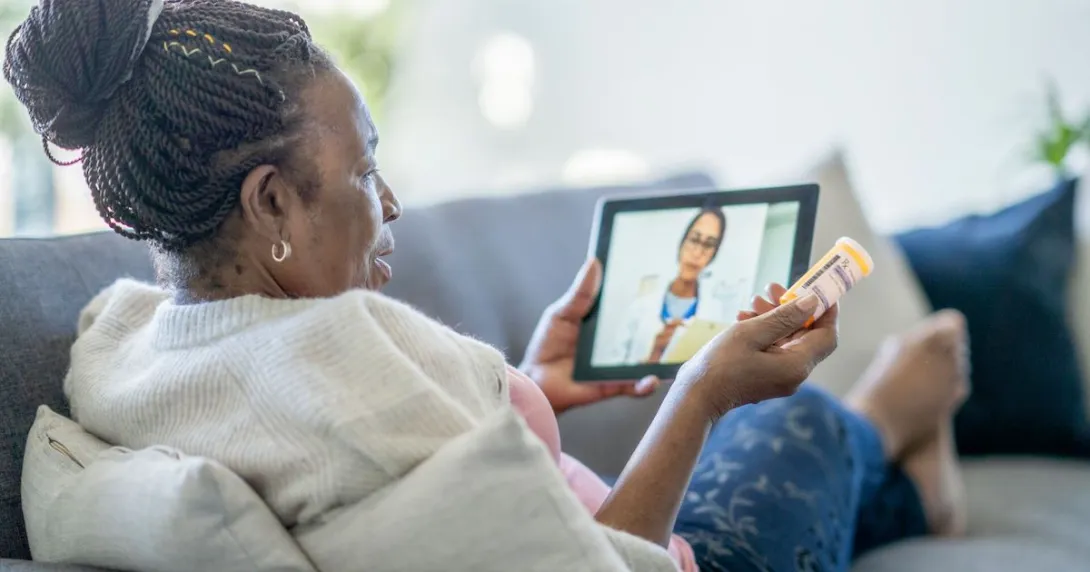 Person sitting on a couch holding a tablet on their lap with a healthcare professional on the screen Person sitting on a couch holding a tablet on their lap with a healthcare professional on the screen