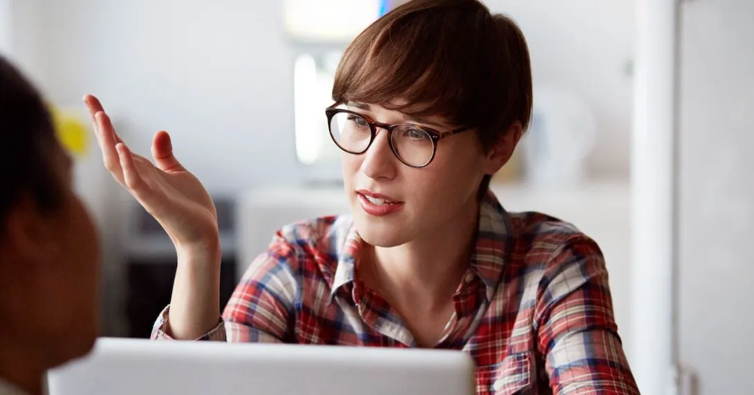 A person with a red plaid shirt on with short hair wearing glass, sitting at a computer with their hand up while talking A person with a red plaid shirt on with short hair wearing glass, sitting at a computer with their hand up while talking
