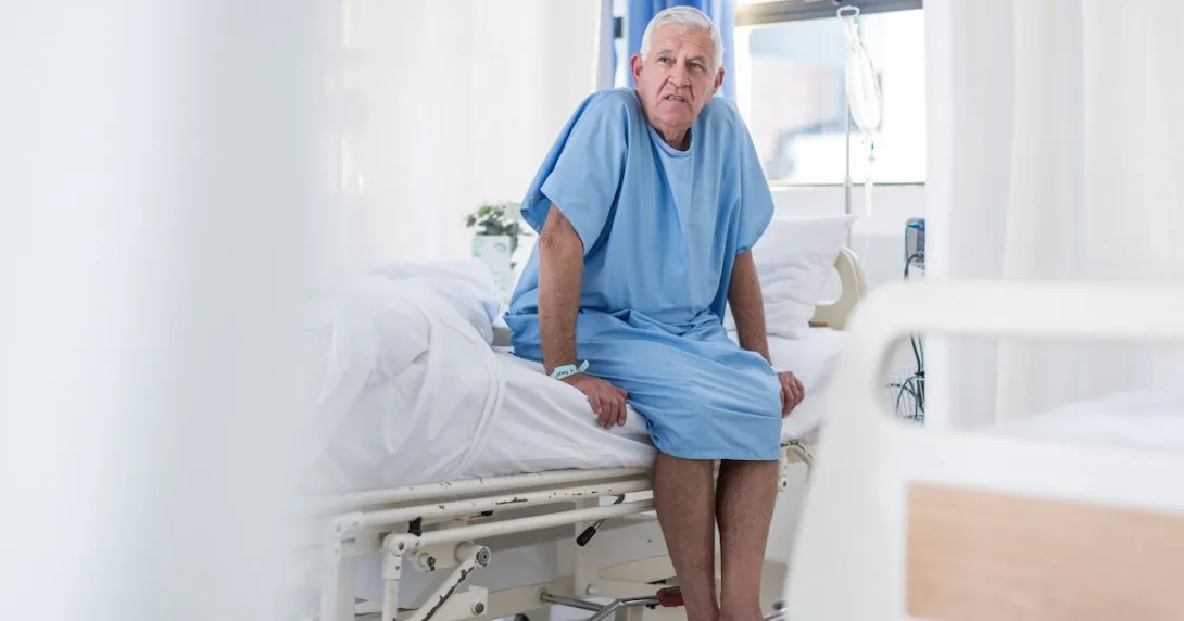 Patient sitting on the side of a bed in a hospital room wearing a hospital gown Patient sitting on the side of a bed in a hospital room wearing a hospital gown