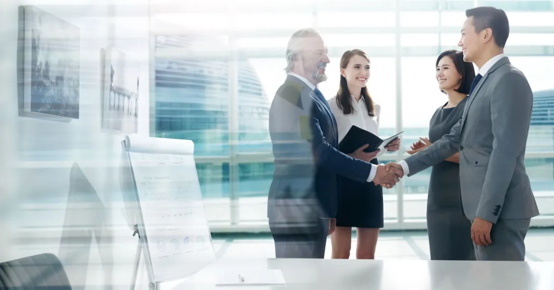 Four people standing in front of a large window with two of them shaking hands and the other two watching Four people standing in front of a large window with two of them shaking hands and the other two watching