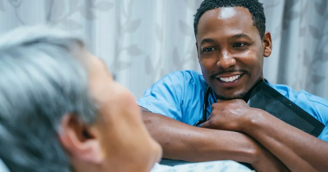 Healthcare provider sitting beside a patient's bed with the patient in it while the provider is holding a tablet Healthcare provider sitting beside a patient's bed with the patient in it while the provider is holding a tablet